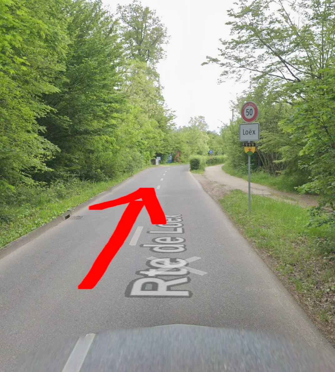 Le panneau Loëx est à droite de la route ; la flèche indique de continuer vers la statue visible plus loin à gauche.